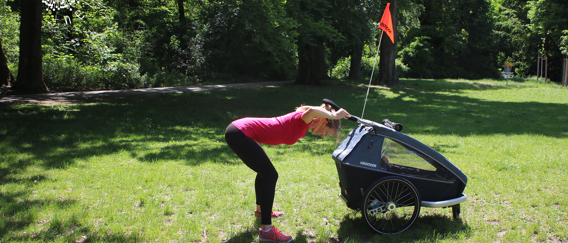 Woman performing push exercise while gripping Croozer bike trailer handlebar with safety flag in park